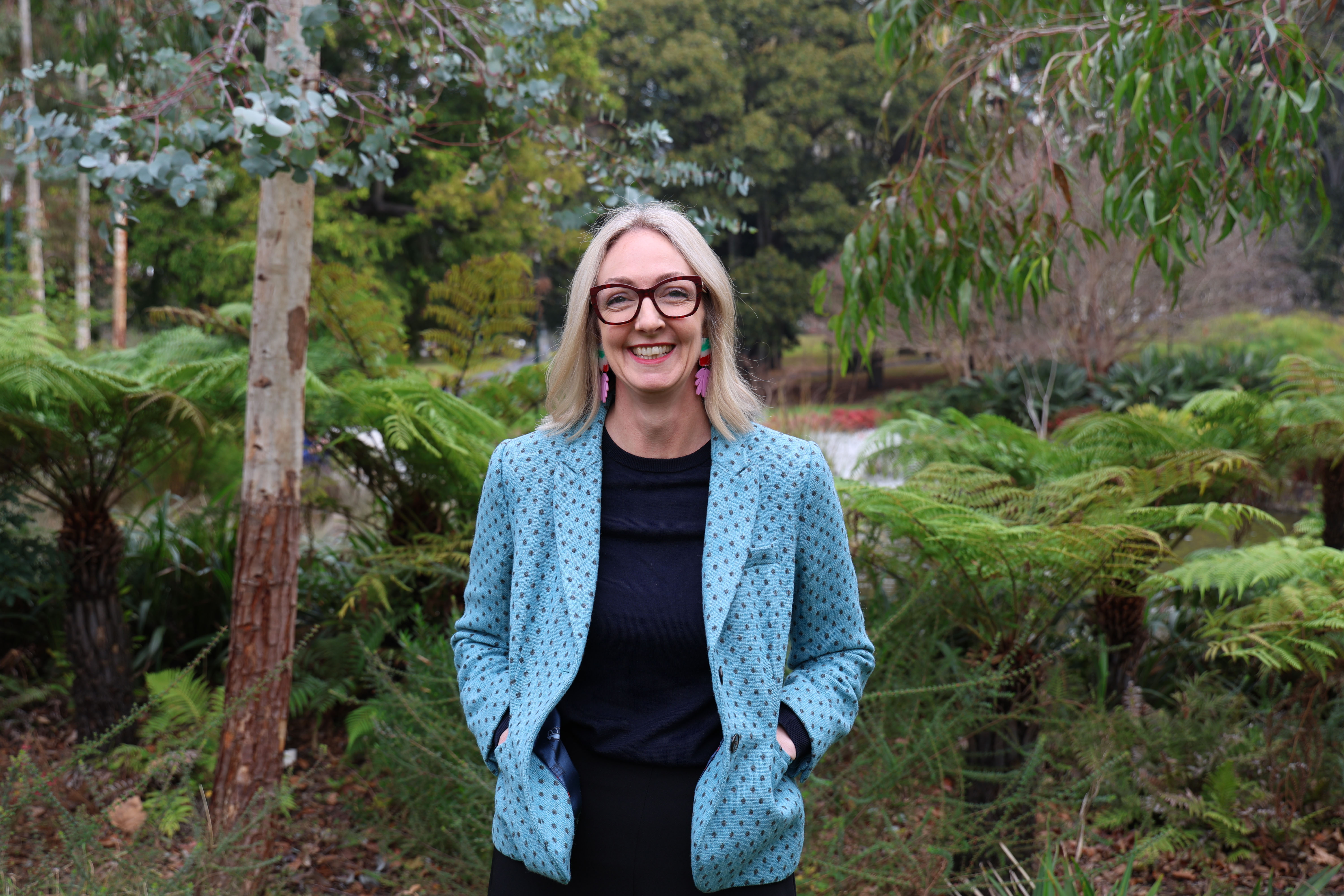 Lisa, dressed in a teal spotted blazer, stands in front of a backdrop of ferns and eucalyptus trees.