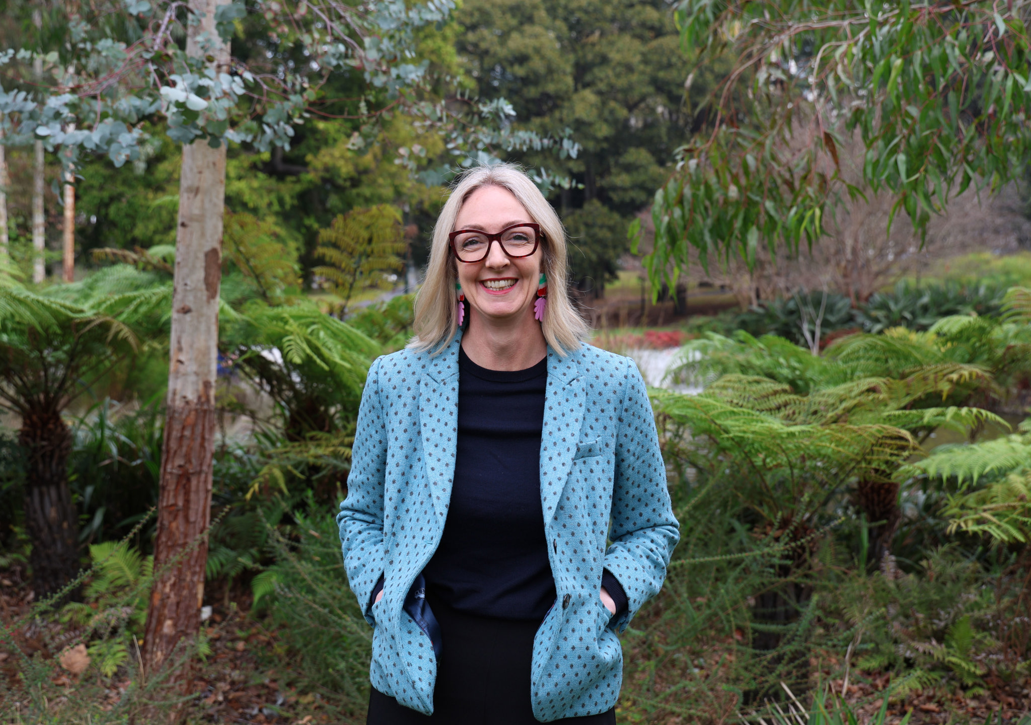 Lisa, dressed in a teal spotted blazer, stands in front of a backdrop of ferns and eucalyptus trees.