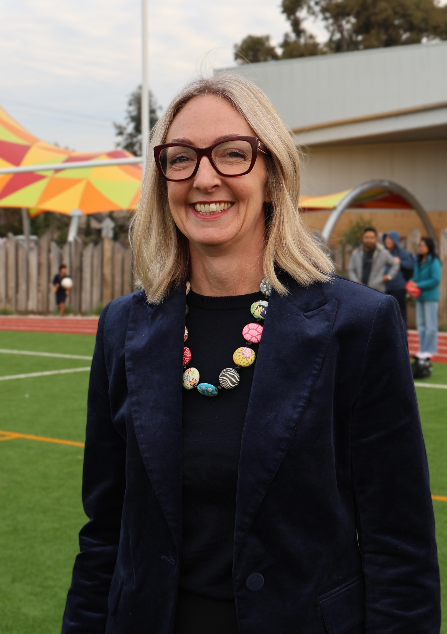 Lisa, wearing glasses and a navy velvet blazer, stands on a sports field at a primary school.